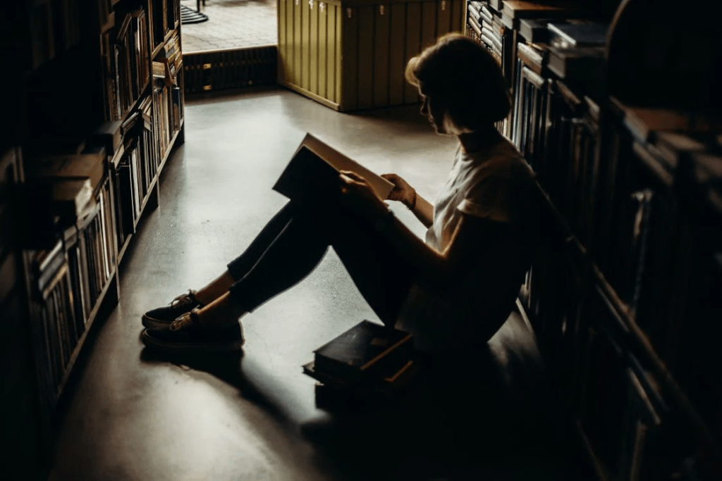 A teen girl sits on the library floor, reading a book. 